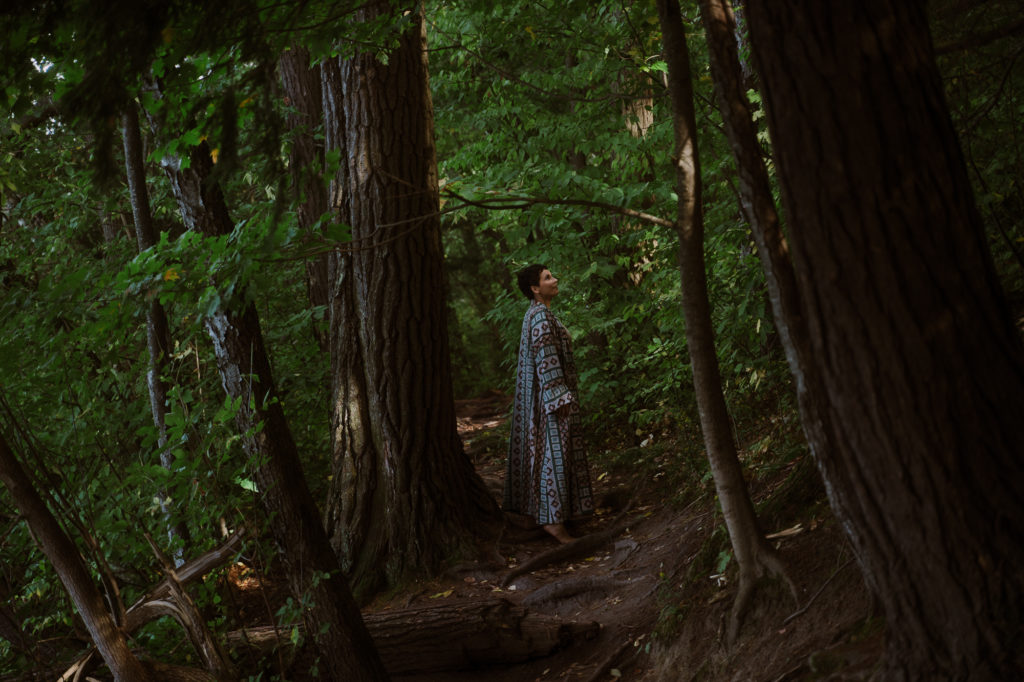 josie walks through forest and looks up at light